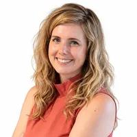 A woman with shoulder-length blonde hair smiles while wearing a sleeveless coral top.