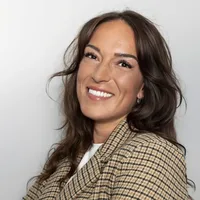 A woman with long brown hair wearing a checked blazer and white top smiles in front of a plain light background.