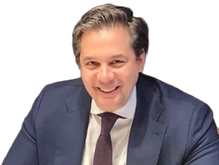 A man in a dark suit, white shirt, and tie is smiling while seated at a table against a plain background.