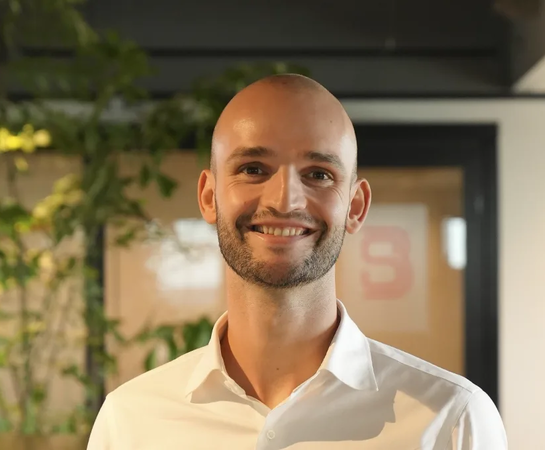 A man with a shaved head and trimmed beard, wearing a white shirt, smiles while standing indoors with plants and a blurred background behind him.