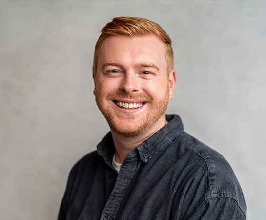 Smiling person with short red hair and a trimmed beard, wearing a dark button-up shirt, posing against a light gray background.