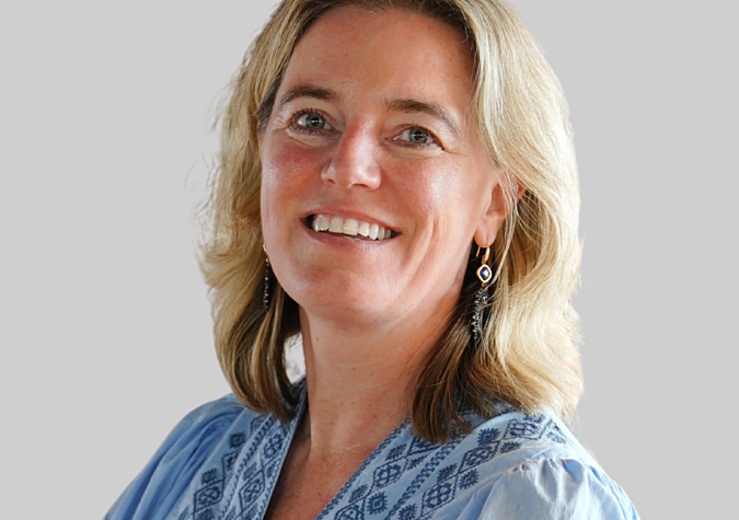 A woman with blonde hair wearing a light blue patterned blouse and earrings smiles at the camera against a plain light background.