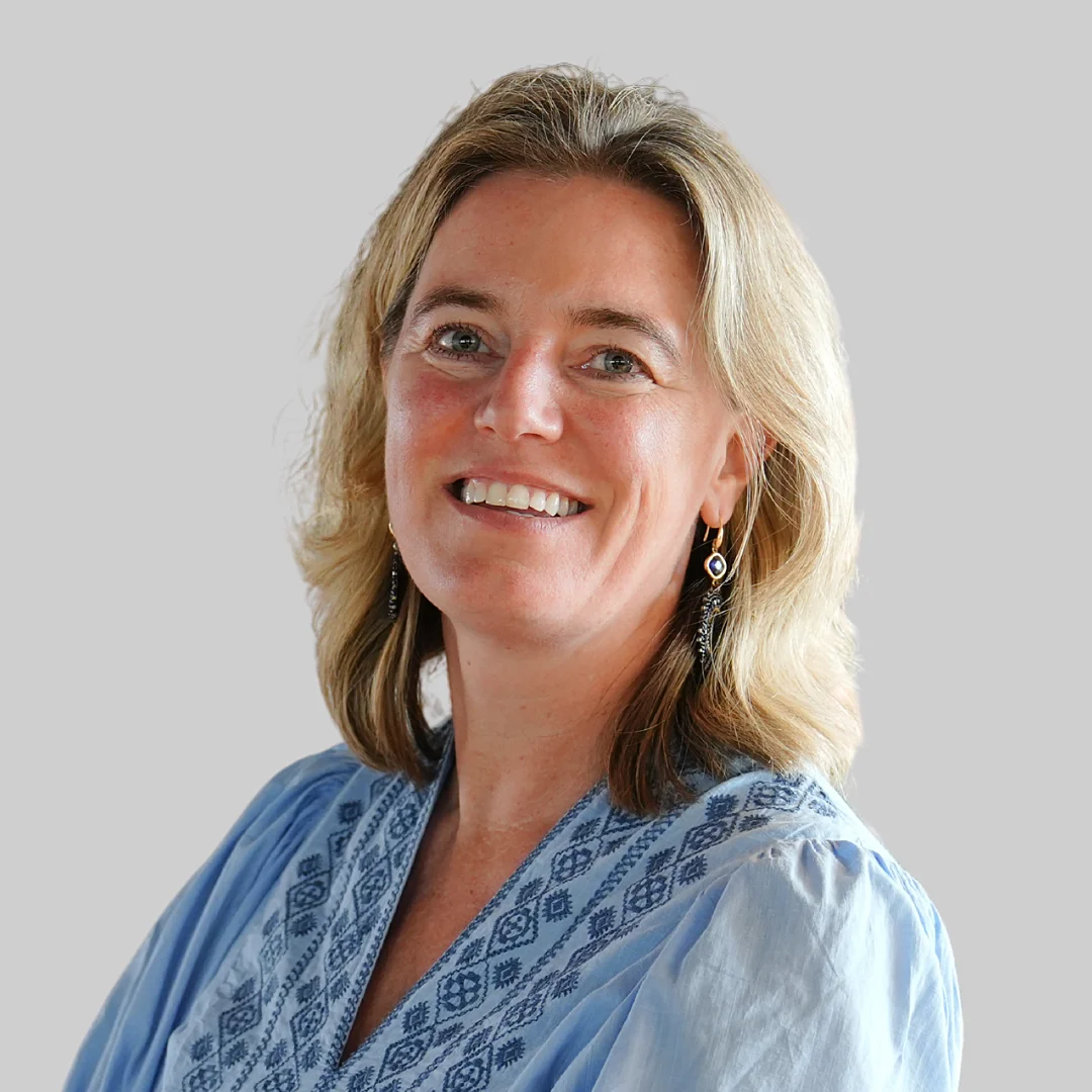 A woman with blonde hair wearing a light blue patterned blouse and earrings smiles at the camera against a plain light background.